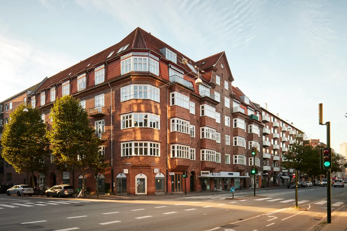 En smuk bygning i mursten med store vinduer og grønne træer ved vejkrydset i en by.