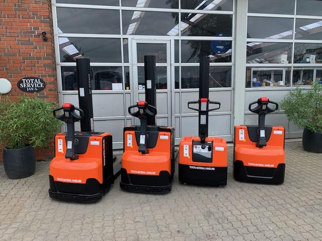 Fire orange gaffeltrucks parkeret foran en bygning med glasdøre og planter i potter.