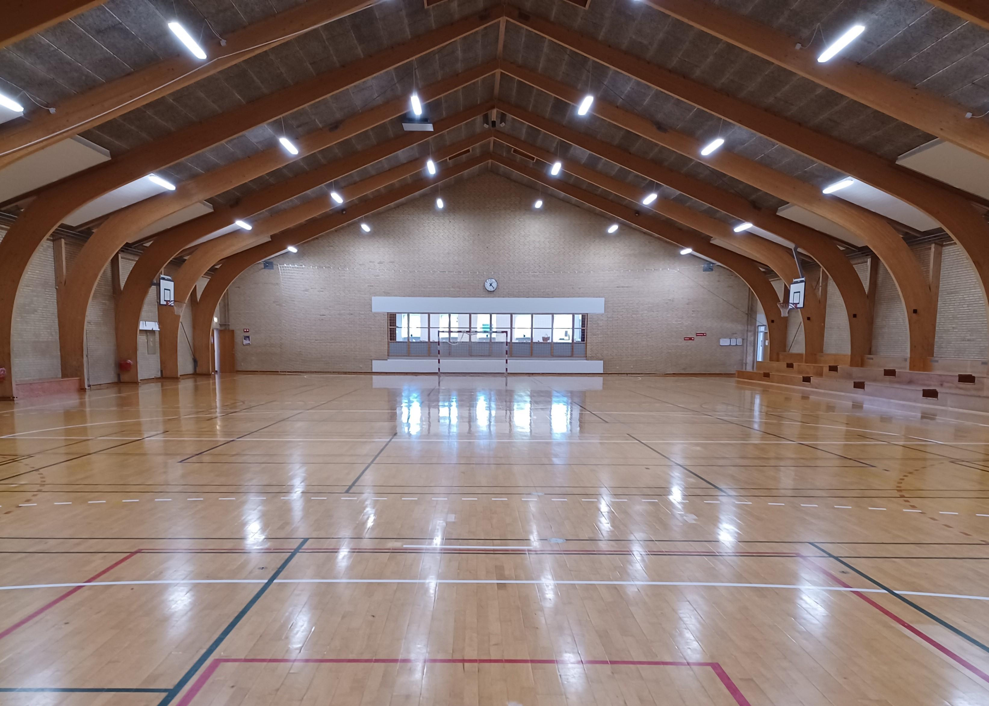 Indendørs sportshal med højt træloft, glat gulv og belysning, klar til aktiviteter og sportsbegivenheder.