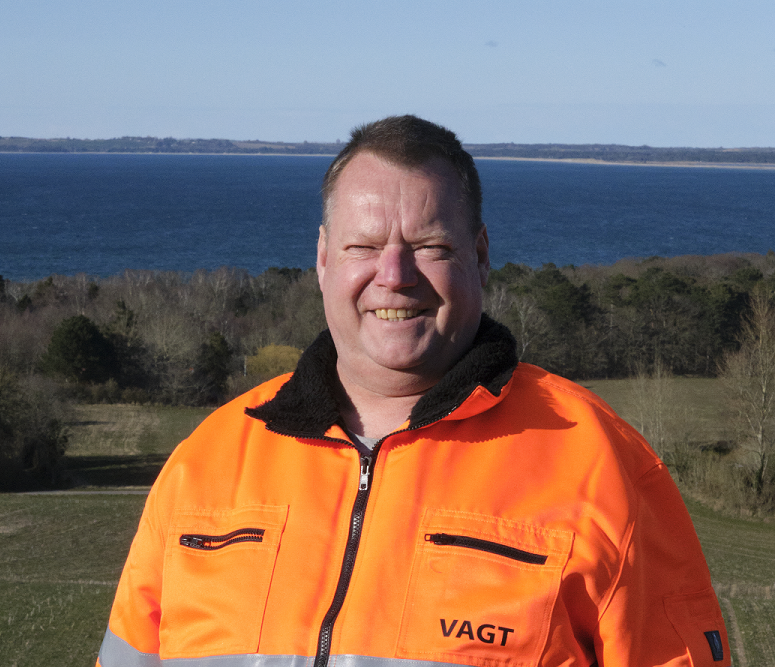 Mand i orange sikkerhedstøj står smilende foran en sø og grønne træer.