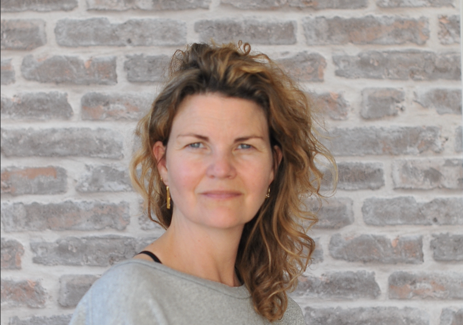 En kvinde med krøllet hår står foran en mur med grå sten. Hun ser direkte ind i kameraet.