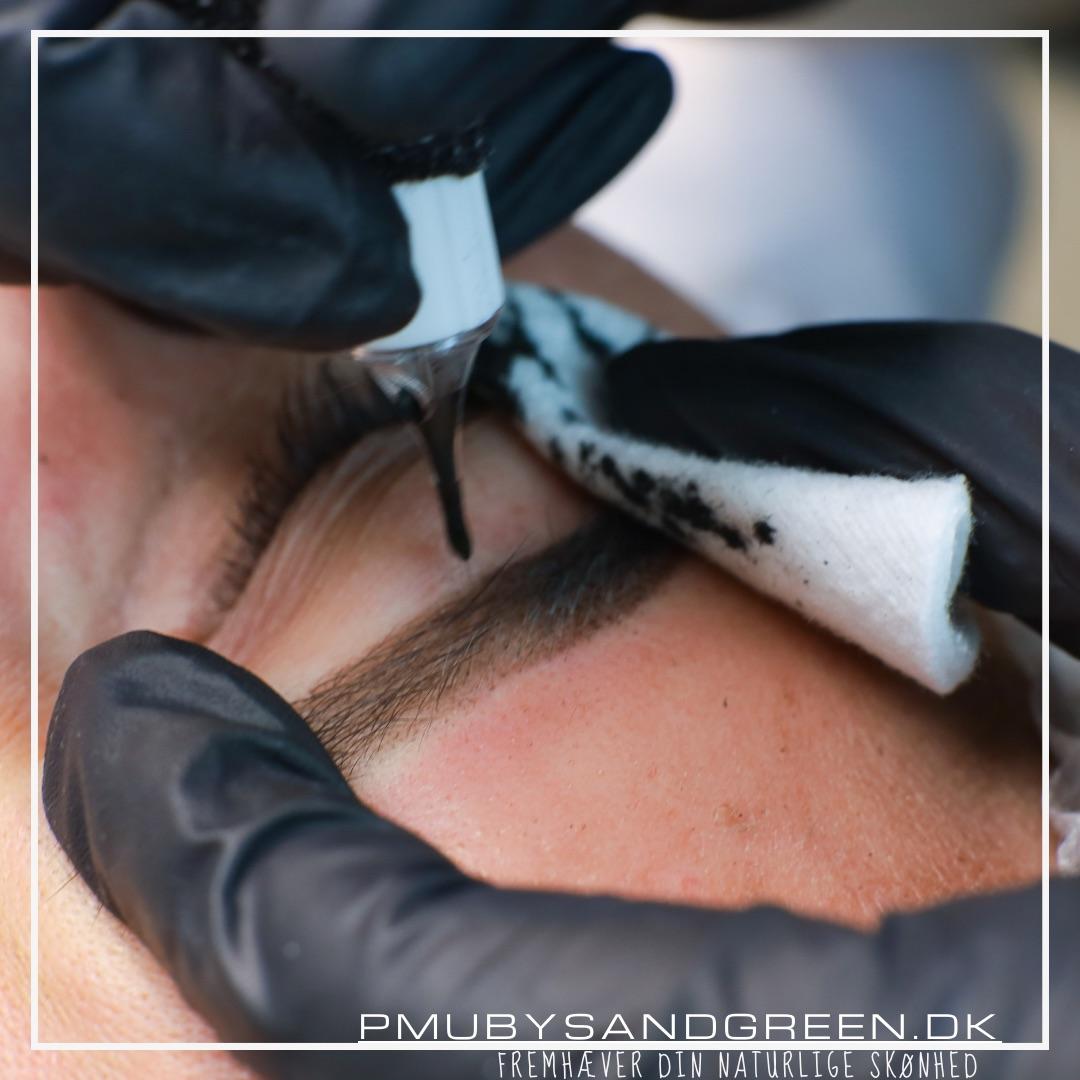 En person får lavet permanent makeup på øjenbrynene med en applikator og beskyttende håndsker.