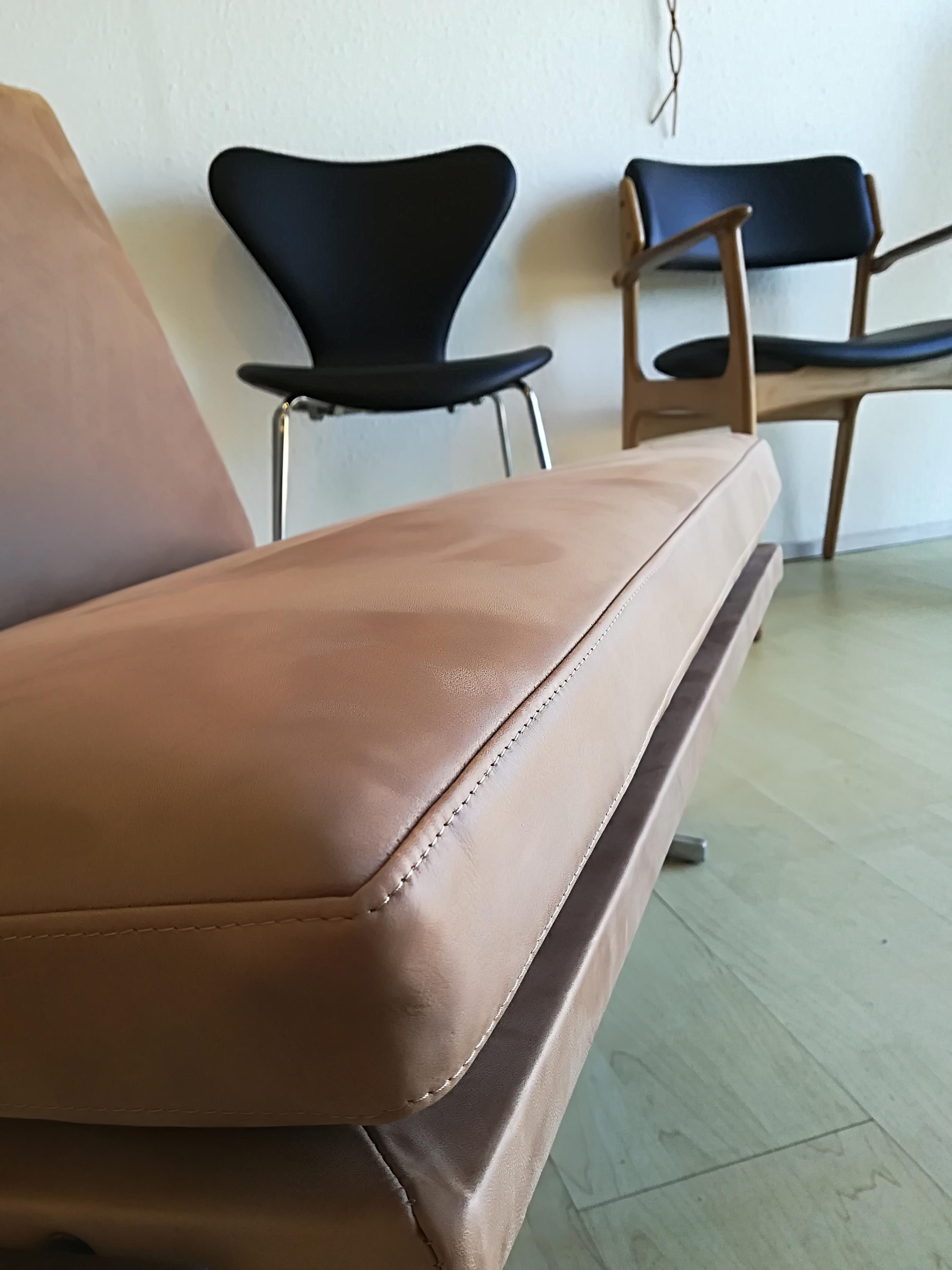 Lædersofa i forgrunden med to stole i baggrunden, en sort og en træstol, på et lyst gulv.