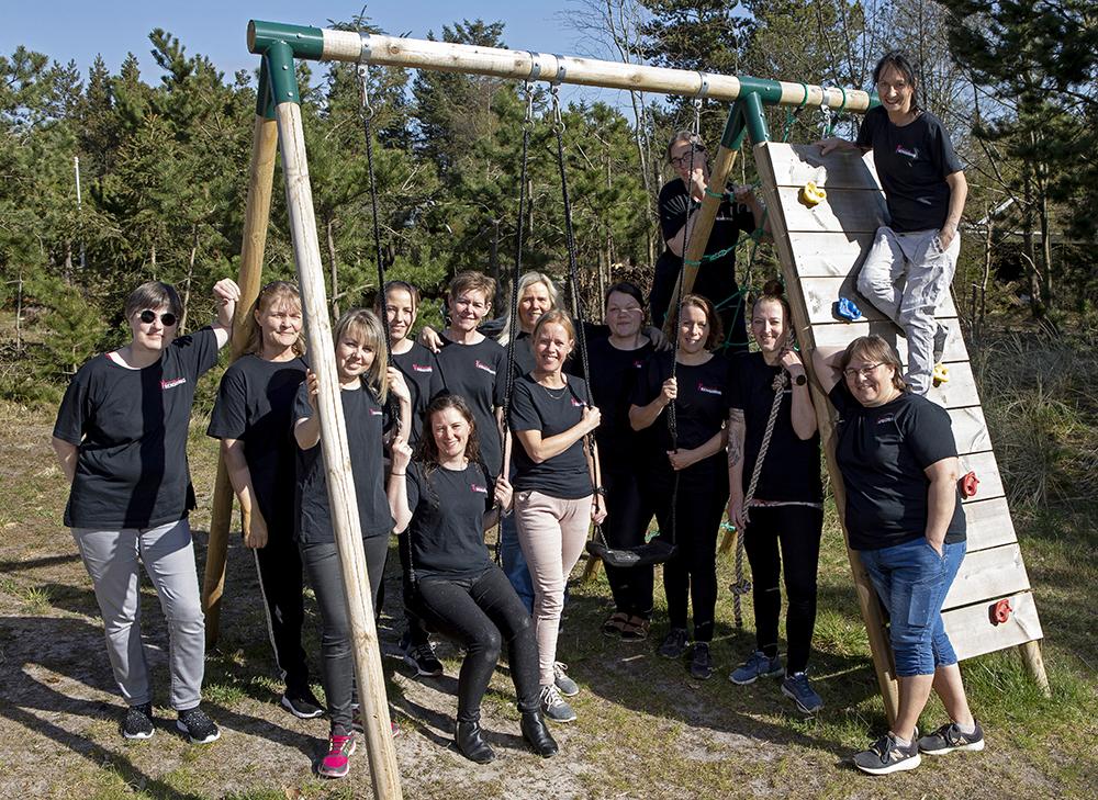 Gruppe af glade mennesker iført sorte t-shirts, der poserer ved en legeplads med gynger og klatrevæg i naturen.