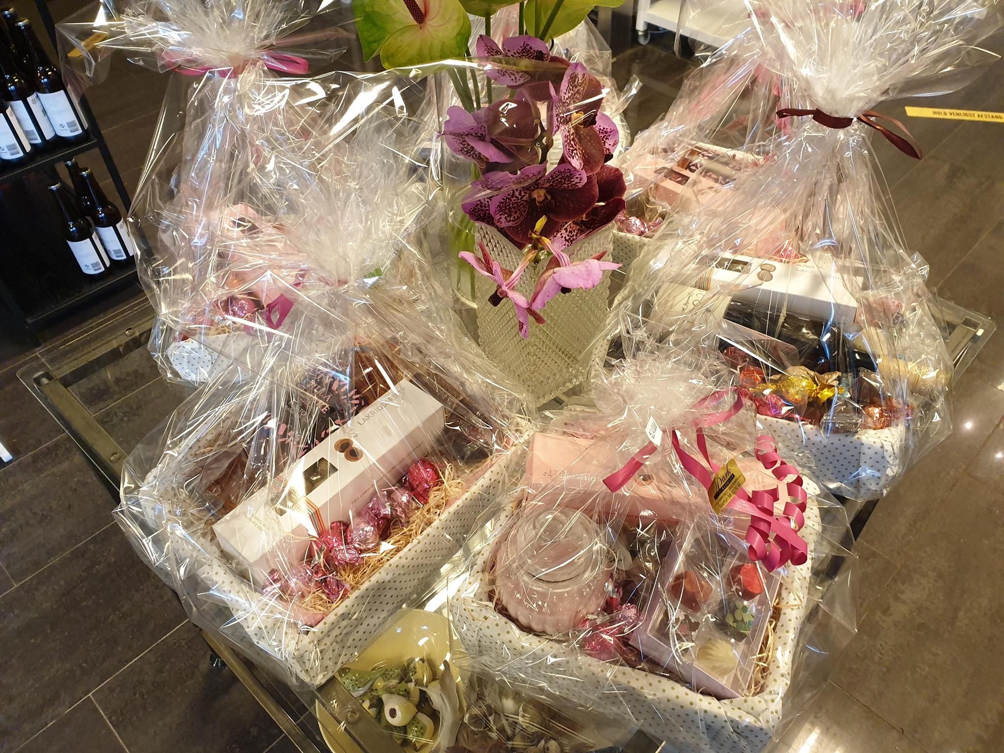 Gavekurve med blomster og lækkerier indpakket i cellofan, præsenteret på et bord.
