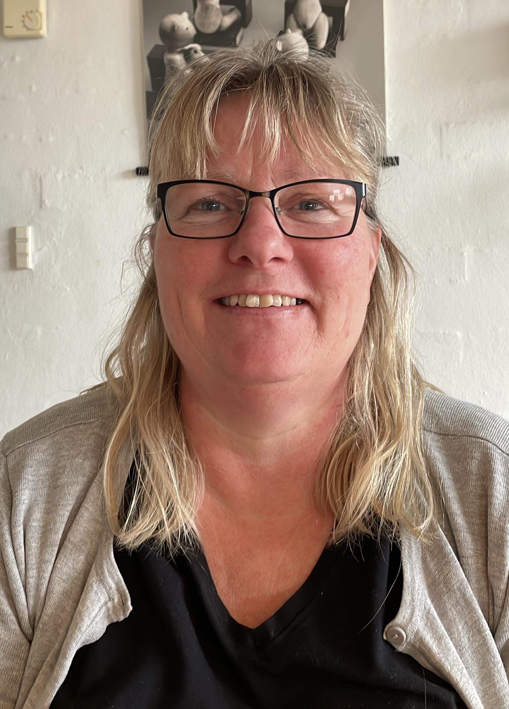 En smilende kvinde med briller og langt hår, iført en grå cardigan, sidder ved et bord i et lyst rum.