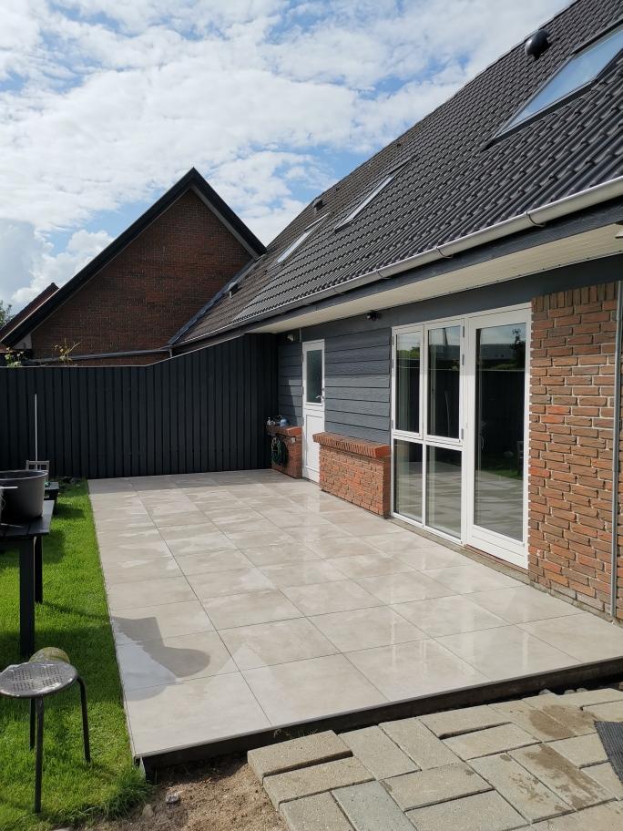 En moderne terrasse med fliser, græsplæne og en bygning i baggrunden under en blå himmel.