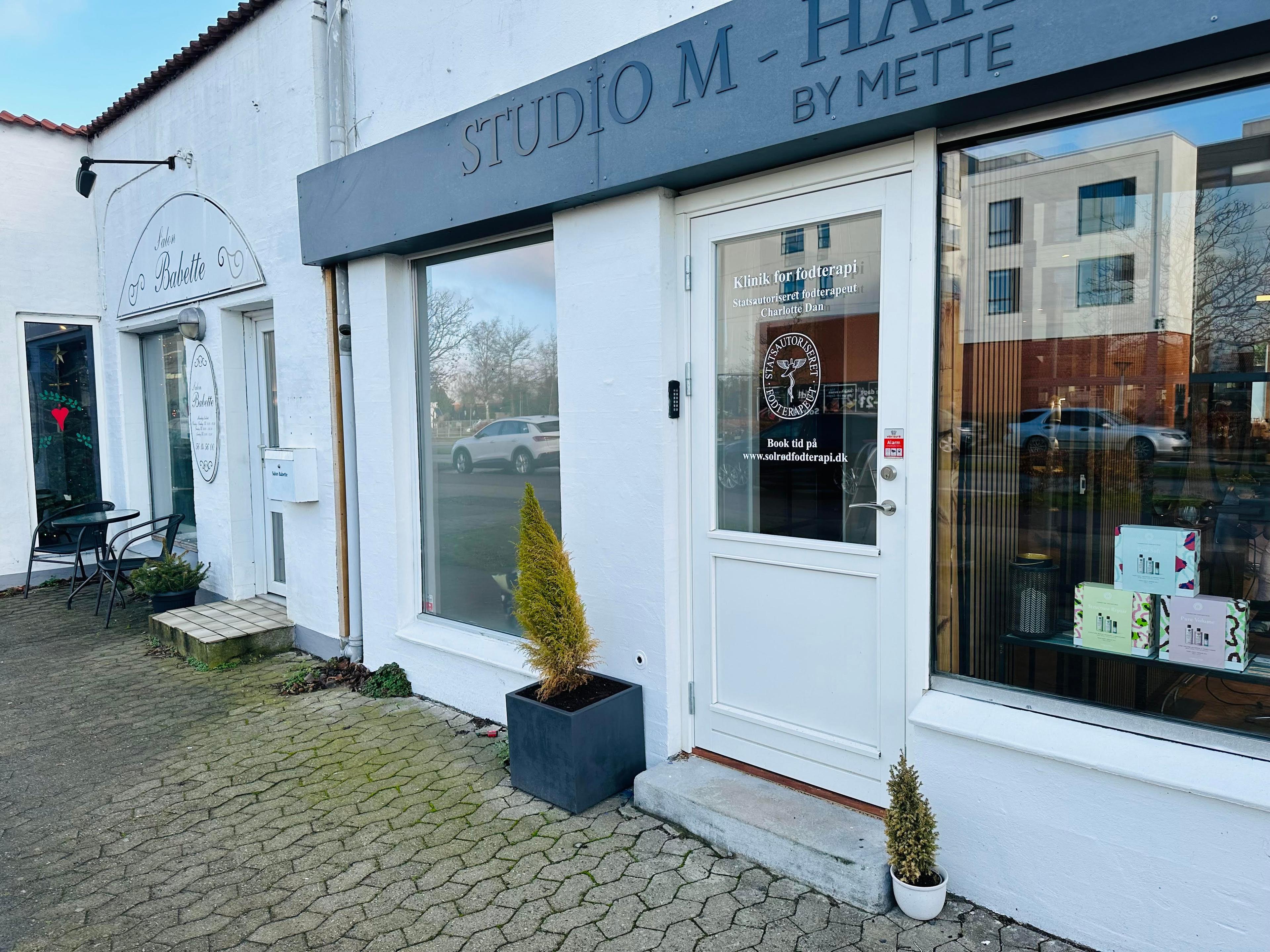 Butiksvindue til Studio M-Hair by Mette med planter og skilte, beliggende på en gade med biler i baggrunden.