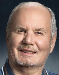 En smilende ældre mand med gråt hår og skæg, iført en blå skjorte.