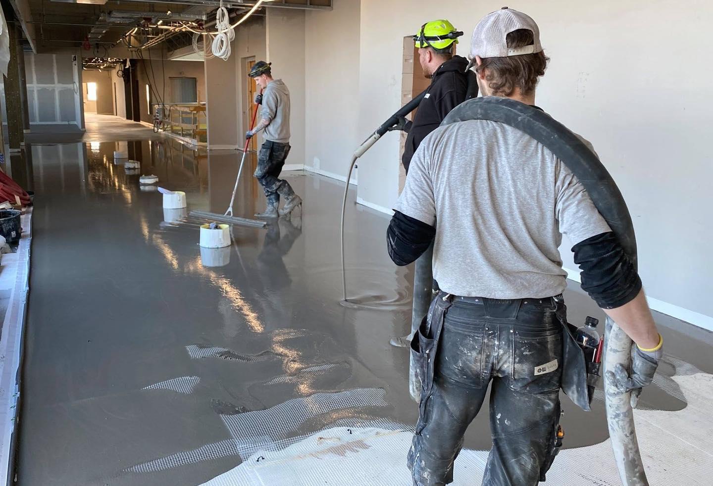Tre håndværkere arbejder med at hælde grå beton i en nybygget gang. Der er malerudstyr og spande omkring dem.