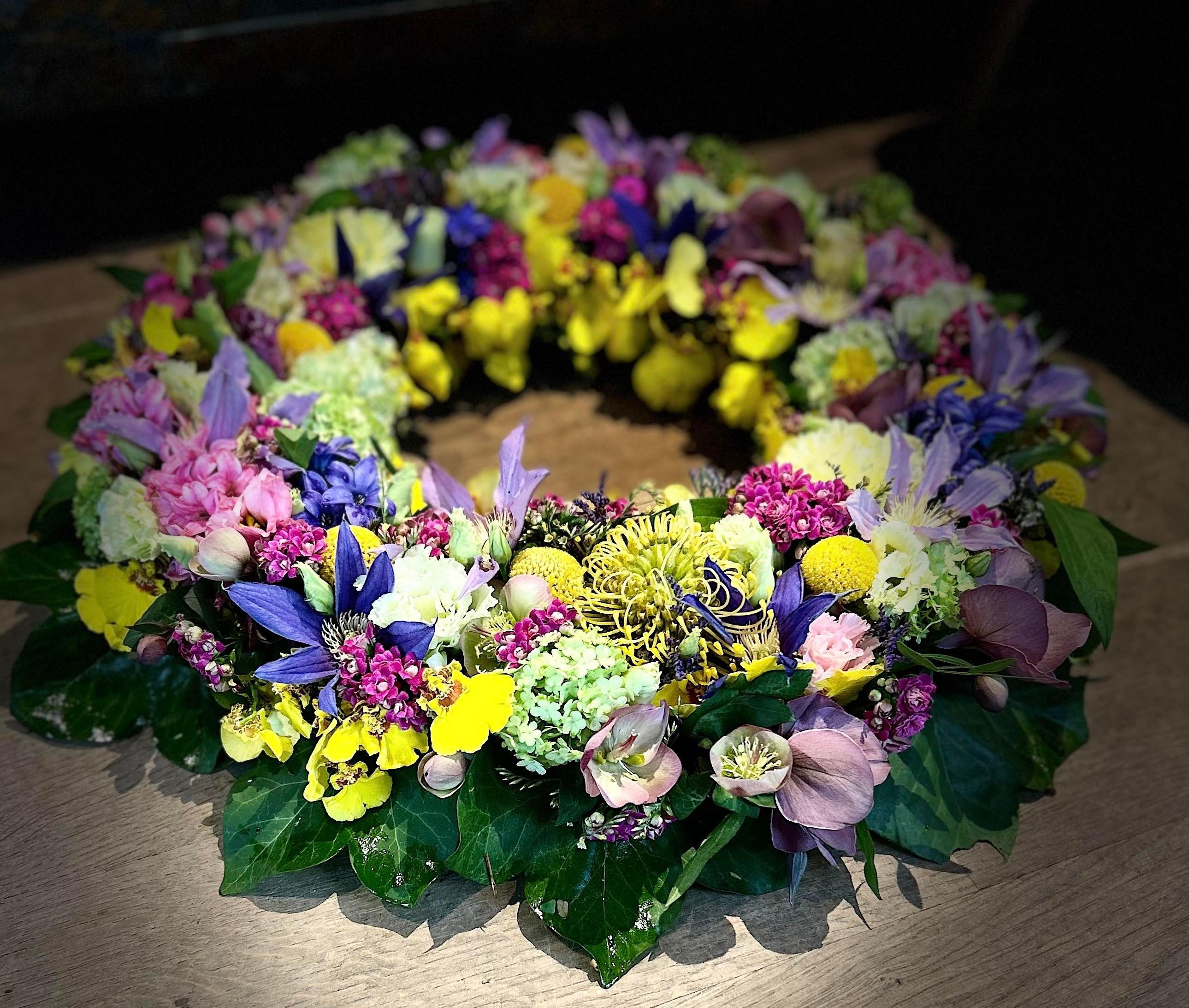 En farverig blomsterkrans med lilla, gule og pink blomster på en træoverflade.