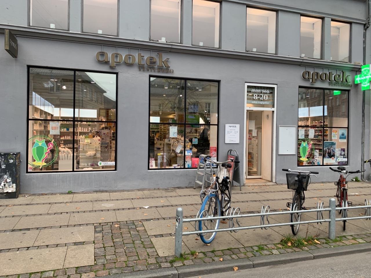Facade af en apotek med store vinduer, cykler parkeret foran og en grøn skiltning.