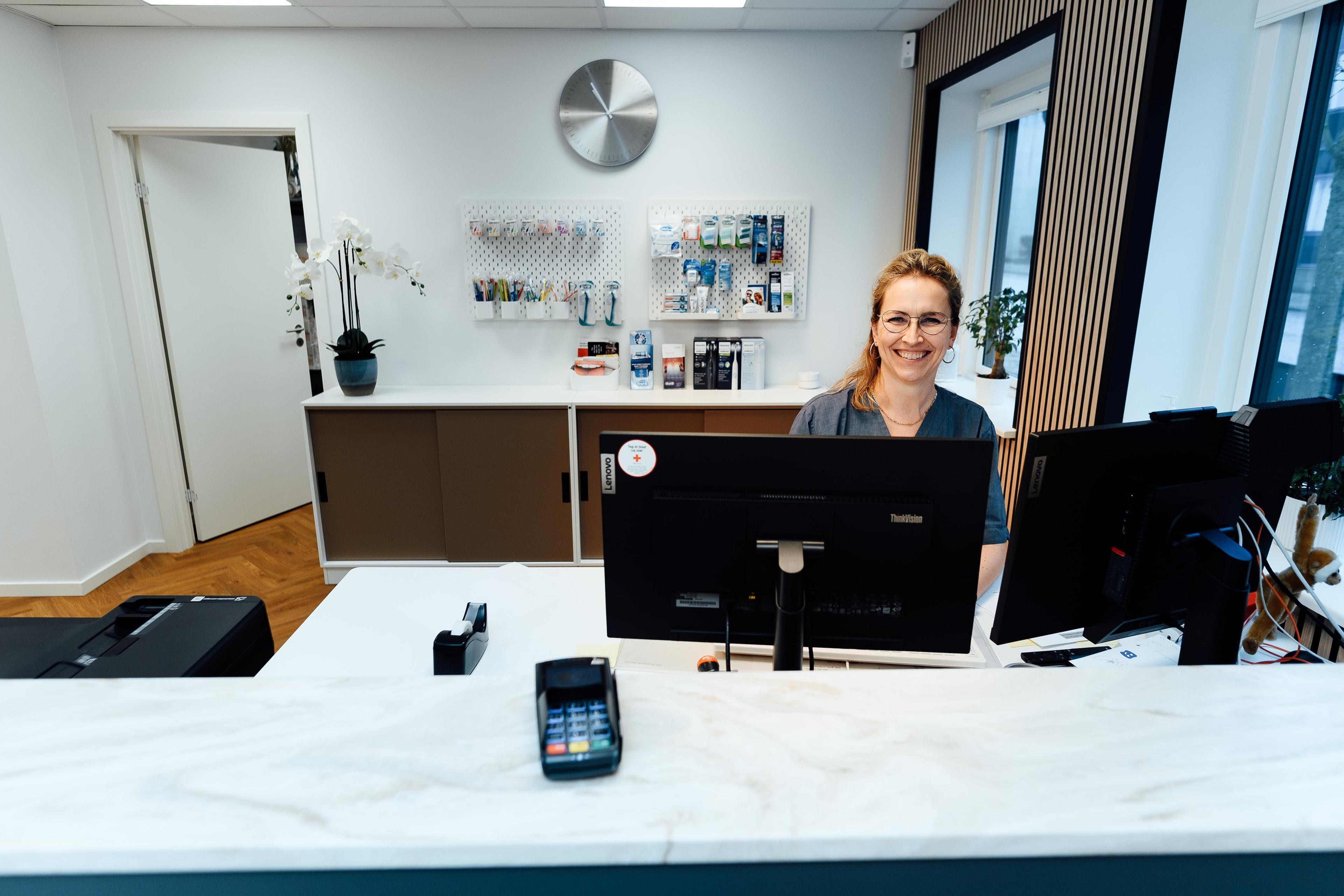 En smilende receptionist sidder ved en computer i en moderne reception med en pæn indretning.