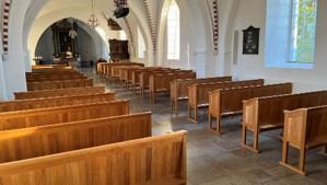 Indvendig udsigt af en kirke med træbænke og store vinduer, der giver naturligt lys.