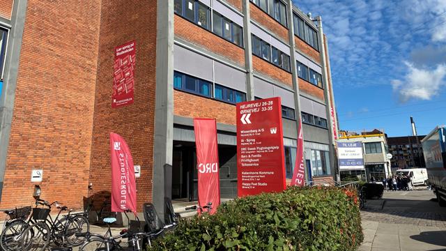 En bygning med rød mursten og store vinduer, omgivet af cykler og flag, der viser information om stedet.
