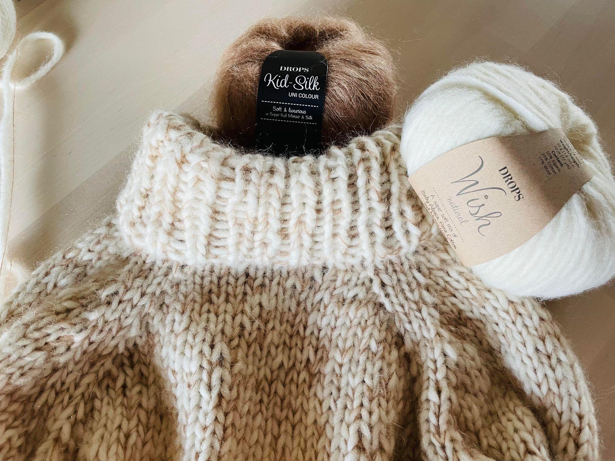 En strikket sweater i beige nuancer med garnnøgler i brunt og hvidt ved siden af.
