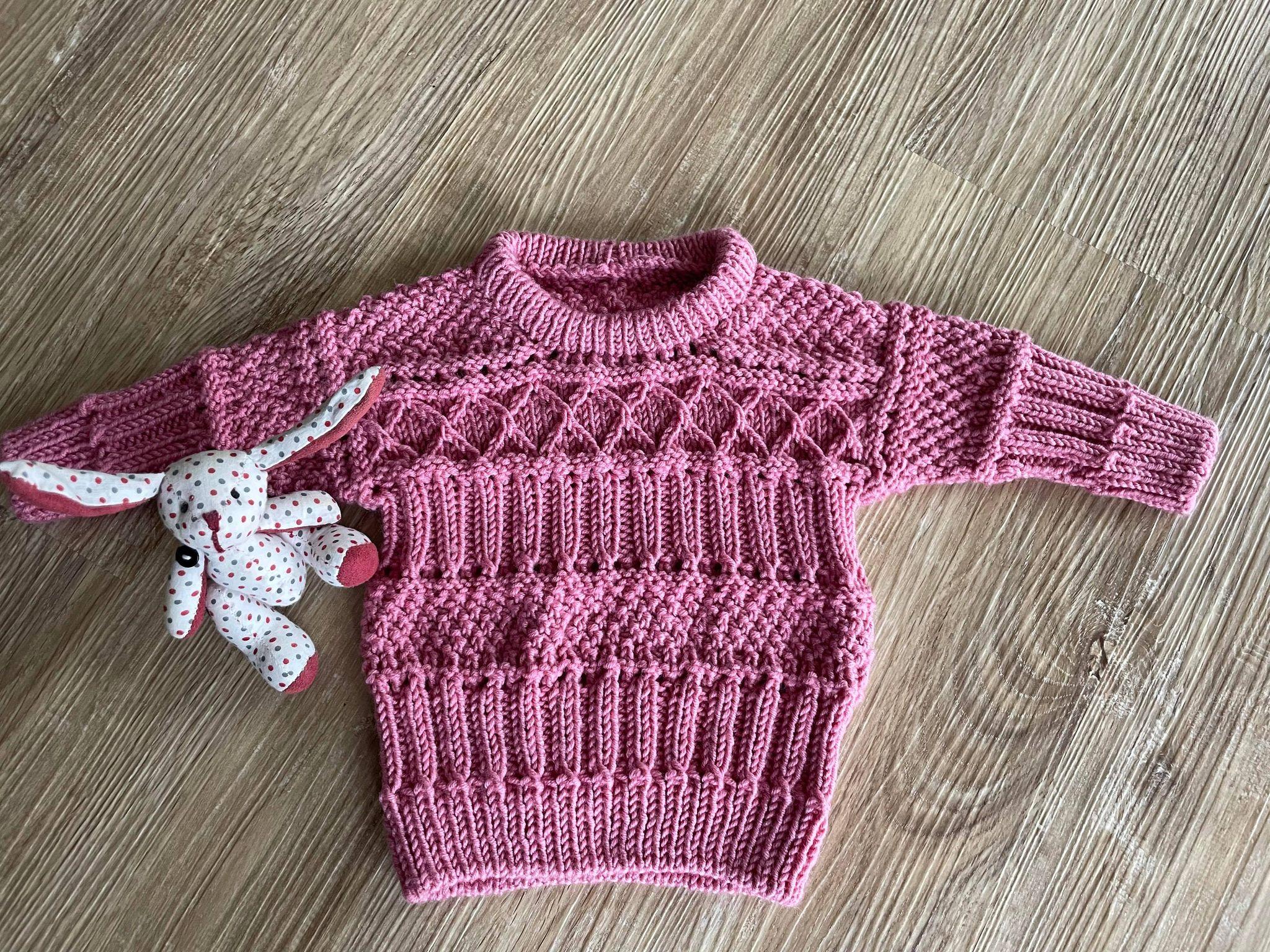En lyserød strikket sweater med mønster og en lille bamse ved siden af på et træbord.