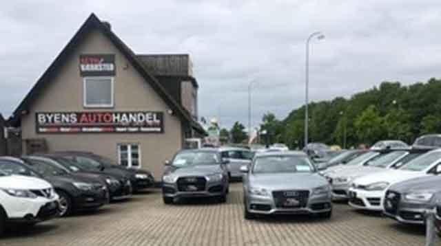 Bilforhandler med flere biler parkeret på en grusplads foran en bygning med skiltet "Byens Auto Handel".