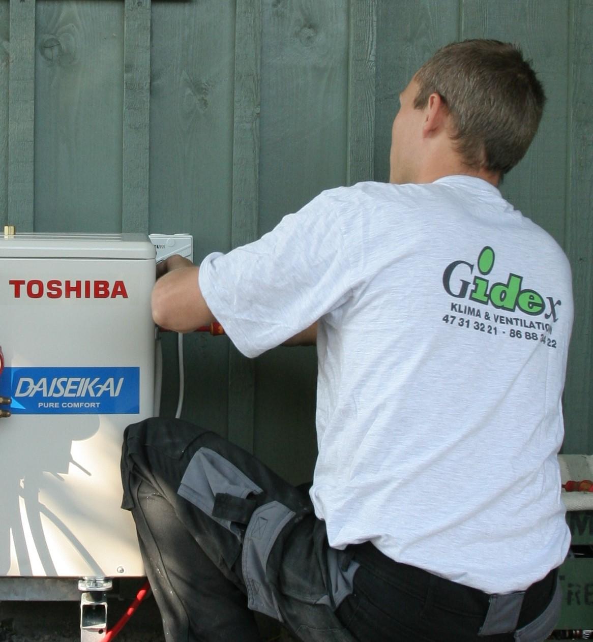 En tekniker installerer en Toshiba klimaanlæg på en væg, iført en hvid T-shirt og arbejdstøj.