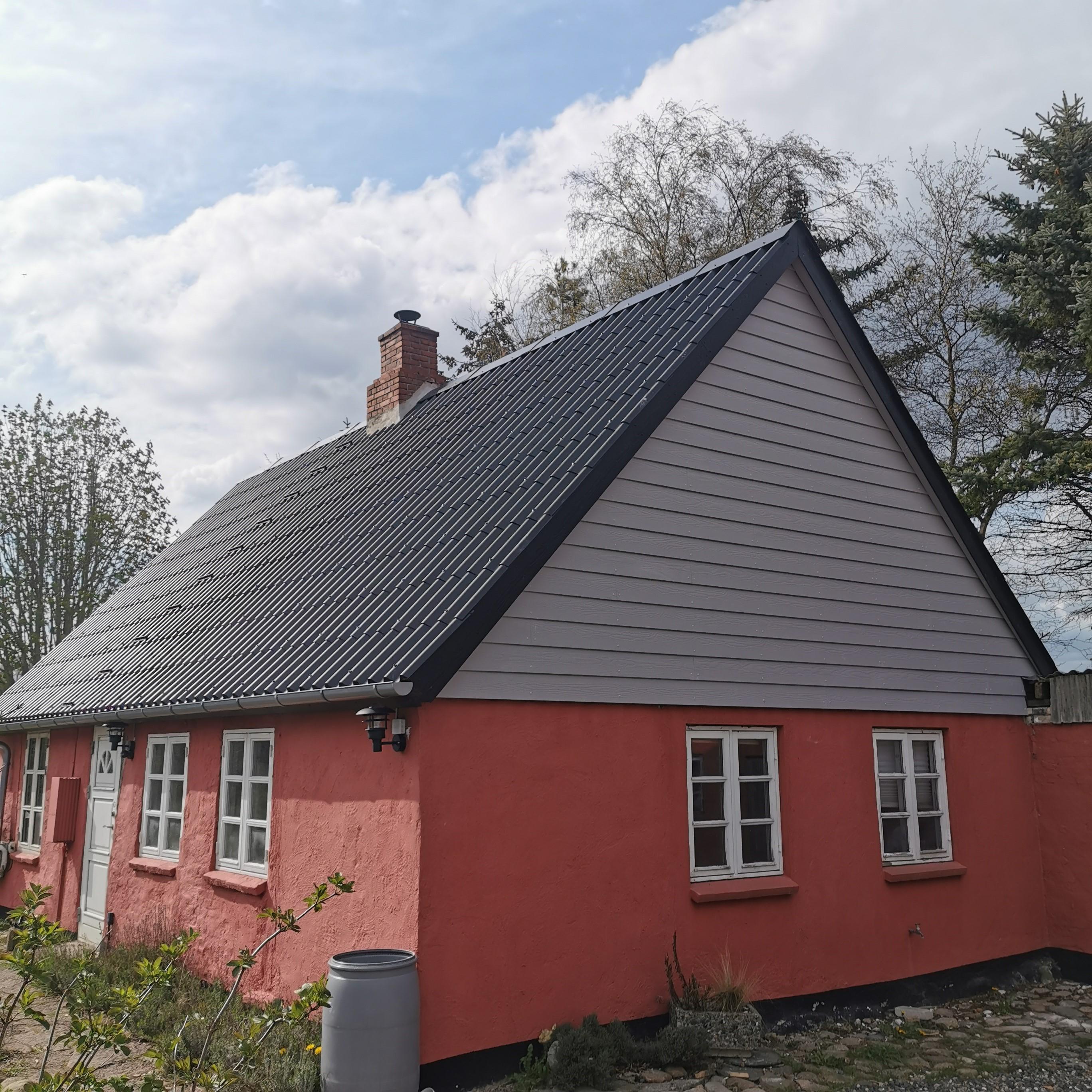 Et charmerende hus med rød facade og grå tag, omgivet af træer og en klar himmel.