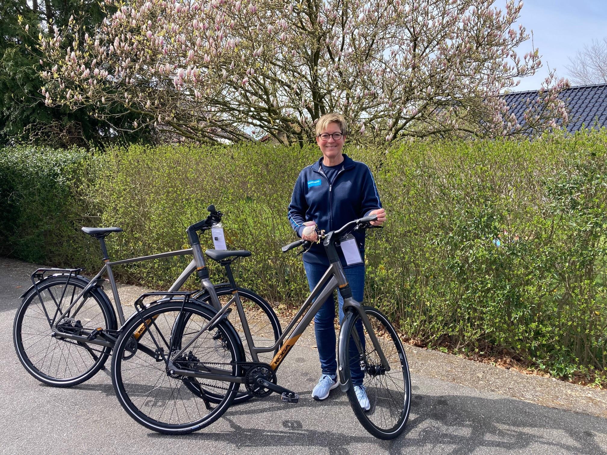 En person står ved to cykler foran en blomstrende busk i et grønt område.