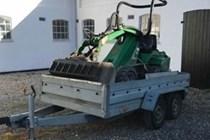 En grøn plæneklipper på en trailer parkeret foran en bygning med grusunderlag.