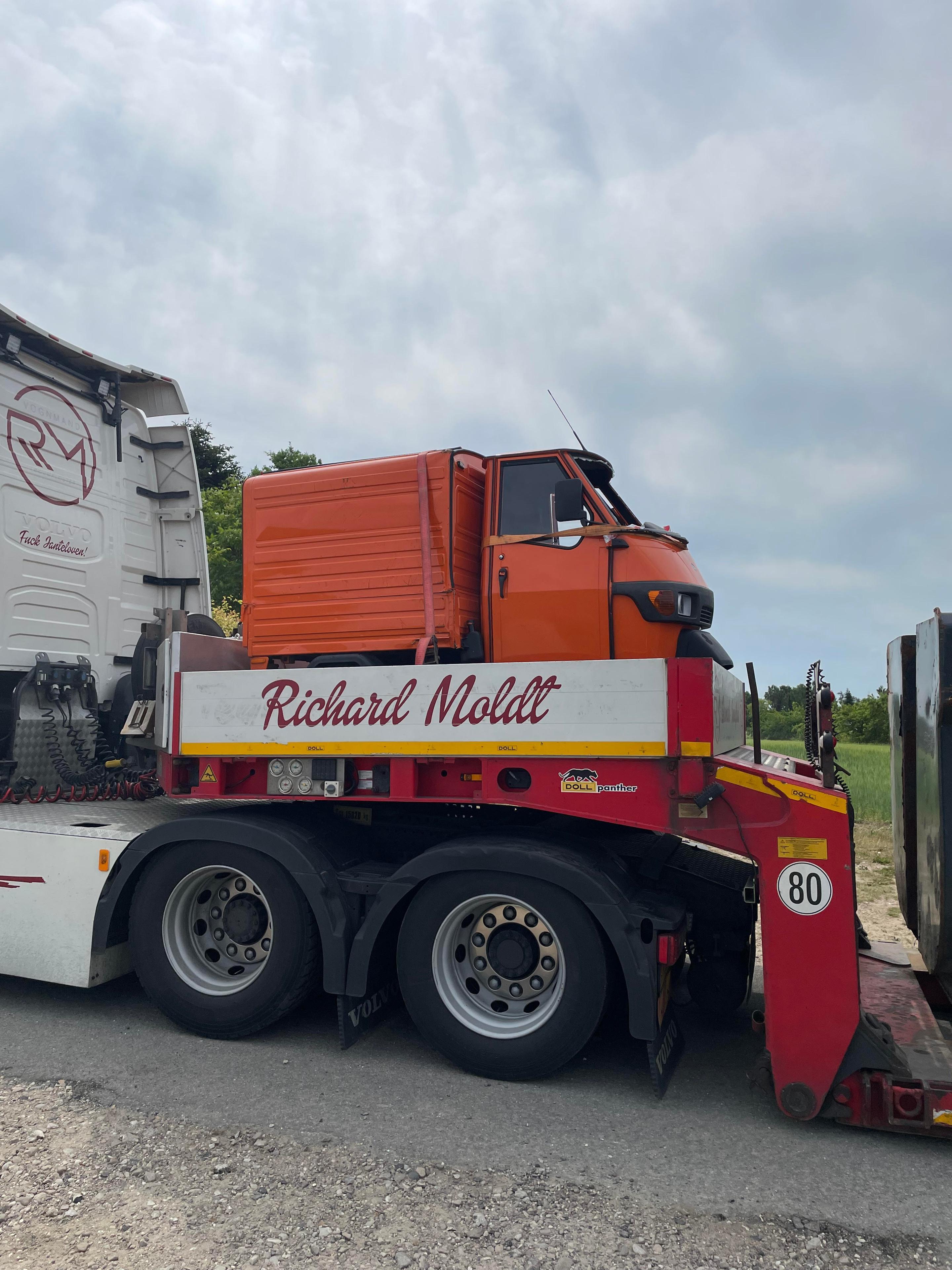 En orange lastbil med teksten "Richard Molt" på siden, parkeret på en grusvej under en skyet himmel.