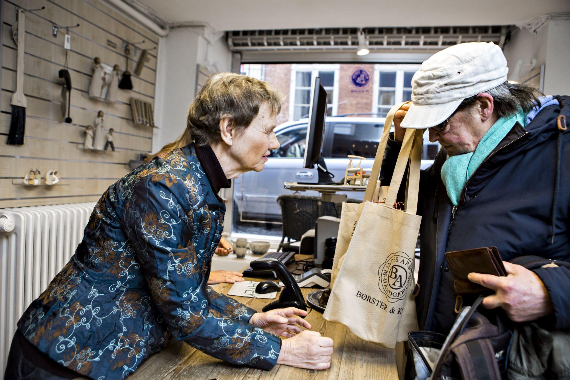 Kunde taler med ekspedient i en butik, mens de står ved en trædisk med en papirpose.