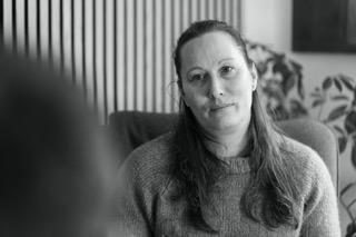 En kvinde med langt hår sidder i en stol og ser opmærksomt på en anden person i et hyggeligt rum.