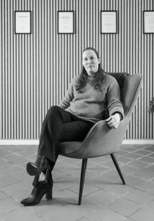 Kvinde sidder i en moderne stol med benene over kors, iført en sweater og bukser, foran en væg med rammede certifikater.