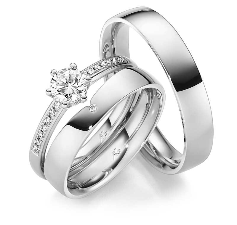 To ringe i sølv med diamanter, herunder en forlovelsesring og en vielsesring, præsenteret i en elegant stak.