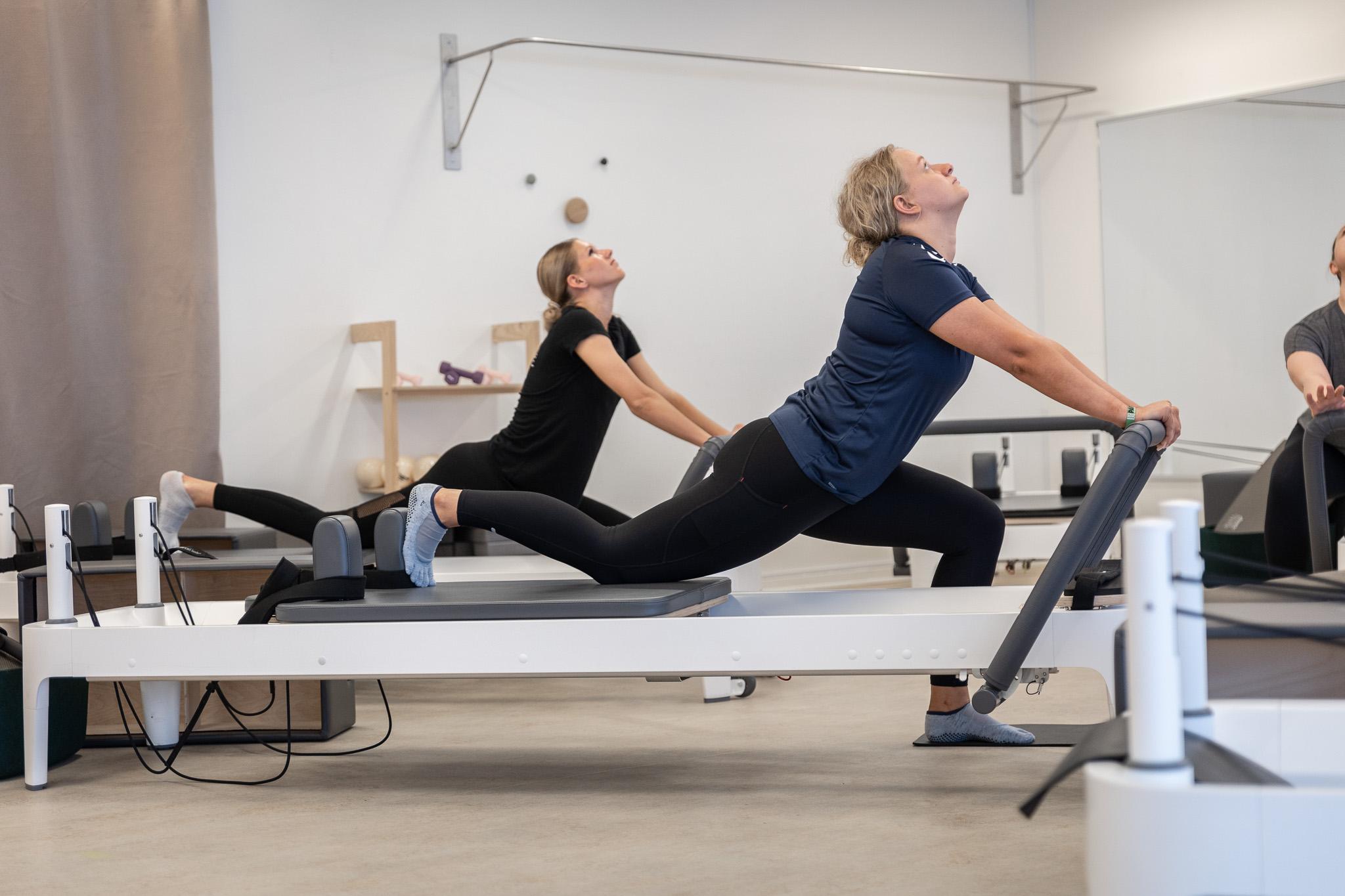 To kvinder, der træner på reformer pilatesmaskiner i et lyst fitnesslokale.