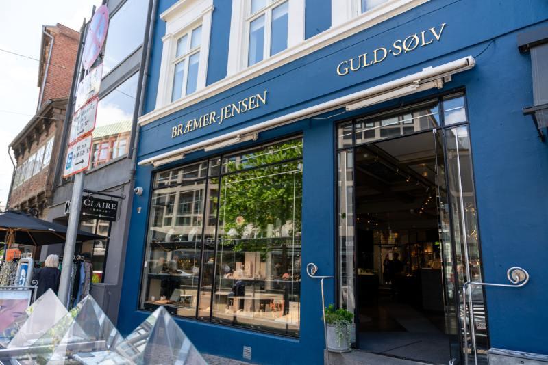 Butiksvindue med navnet "Guld-Solv" på en blå væg, omgivet af træer og mennesker på gaden.