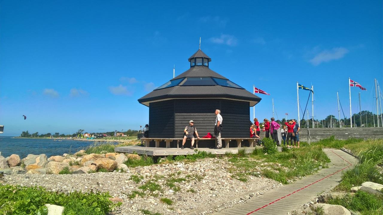 En rund, sort bygning ved vandet med mennesker, der går forbi og flag, der blafrer i vinden.