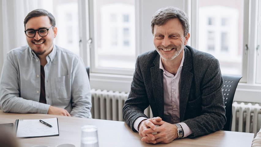 To mænd sidder ved et bord i et mødelokale, smilende og engagerede i en samtale.