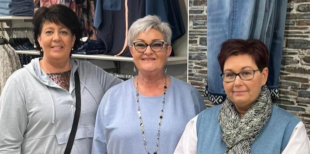 Tre kvinder står sammen i en tøjbutik, iført afslappet tøj og smilende til kameraet.