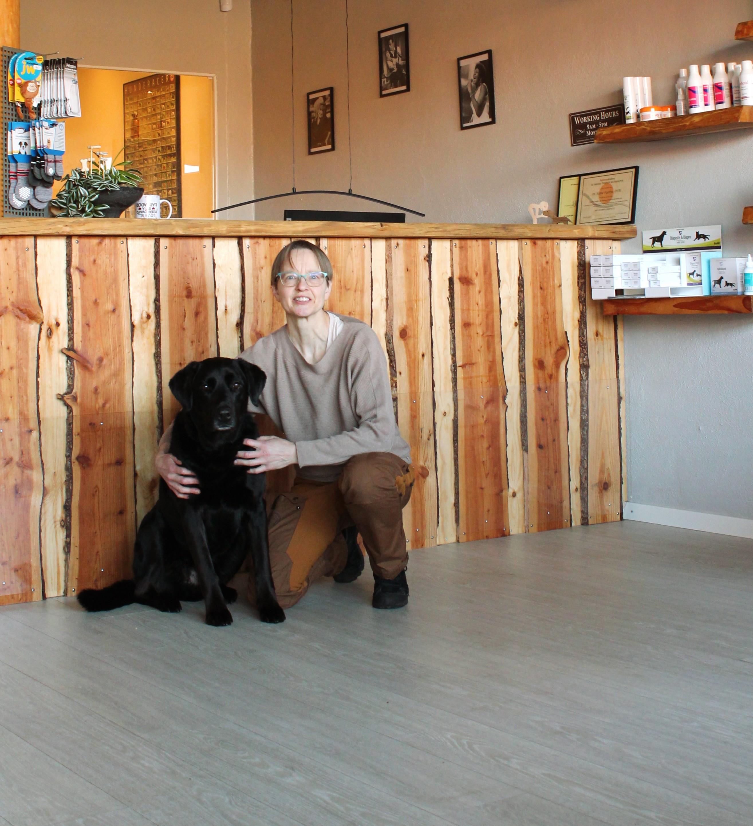 En kvinde sidder på gulvet ved en reception med en sort labradorhund. Baggrunden viser trævægge og hylder.