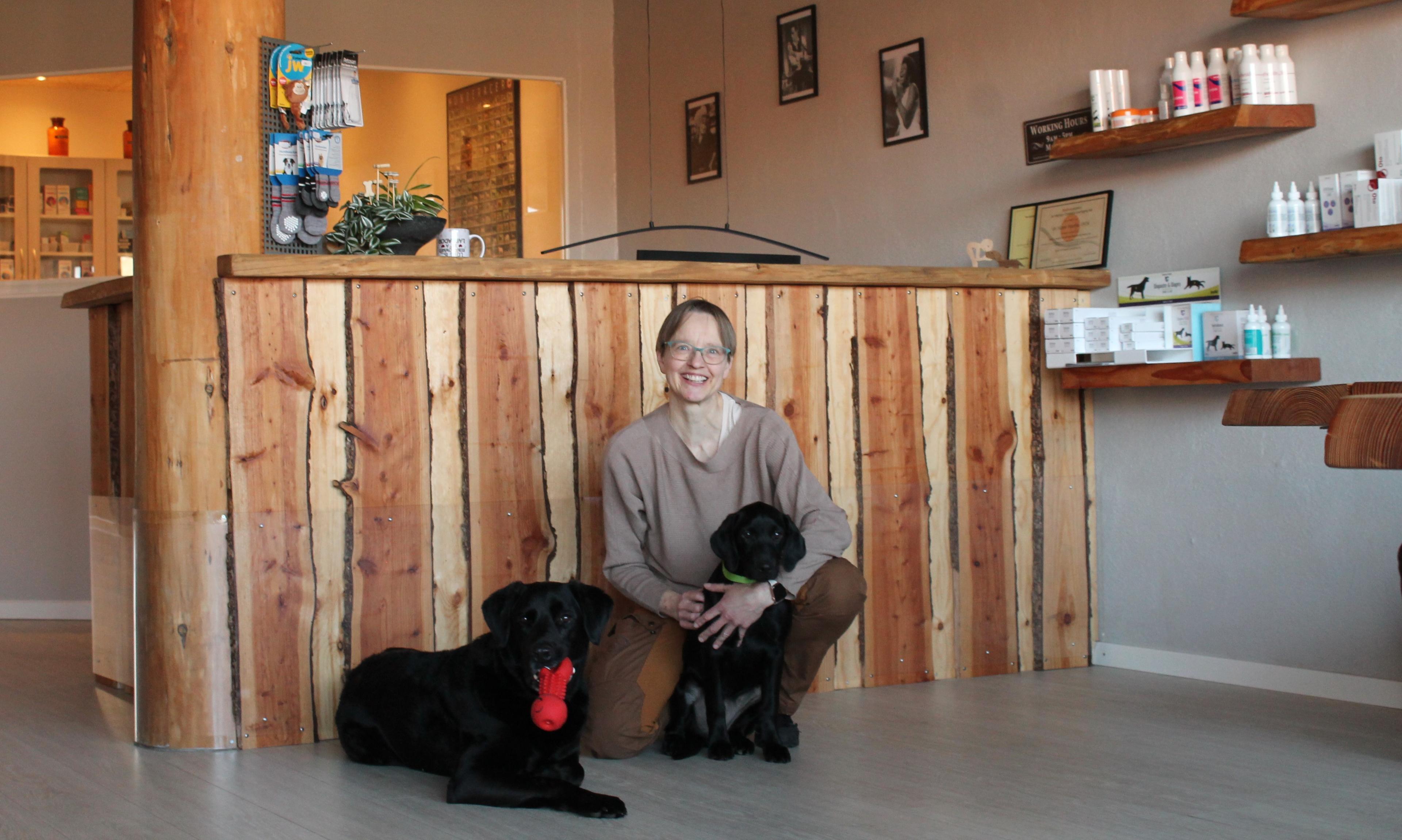 En kvinde sidder på gulvet i en hundesalon med to sorte labradorer ved sin side. Baggrunden viser en trædisk og produkter til hunde.