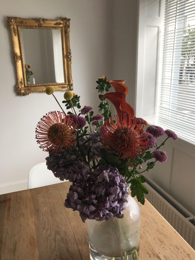 En farverig buket blomster i en glasvase på et træbord med et spejl i baggrunden.