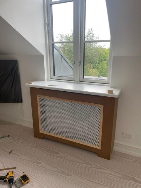 Radiatorafskærmning i træ med hvid topplade ved vindue i moderne rum.