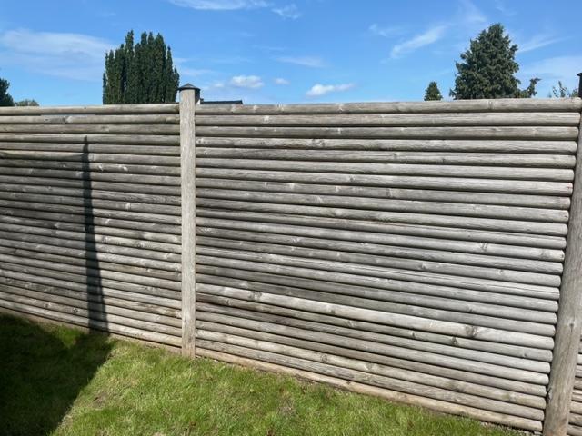 Et gråt træhegn med lodrette planker, omgivet af grøn græsplæne og træer i baggrunden.