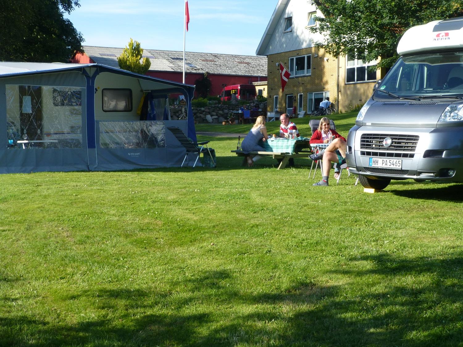 Campingplads med campingvogn, telt og mennesker, der nyder solen på græsset ved en bygning.