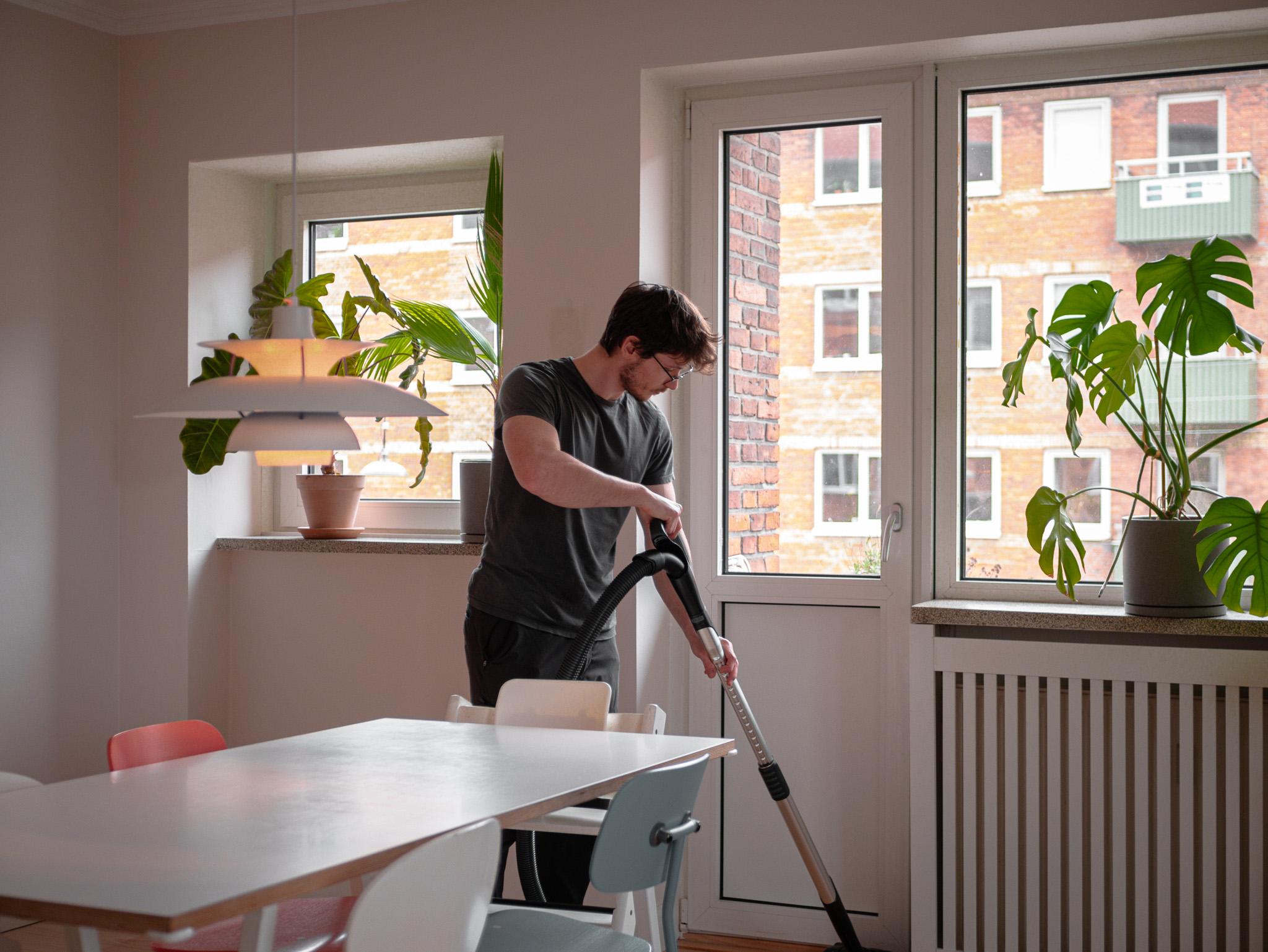 En mand støvsuger i et lyst rum med planter og vinduer, der vender ud mod en bygning.