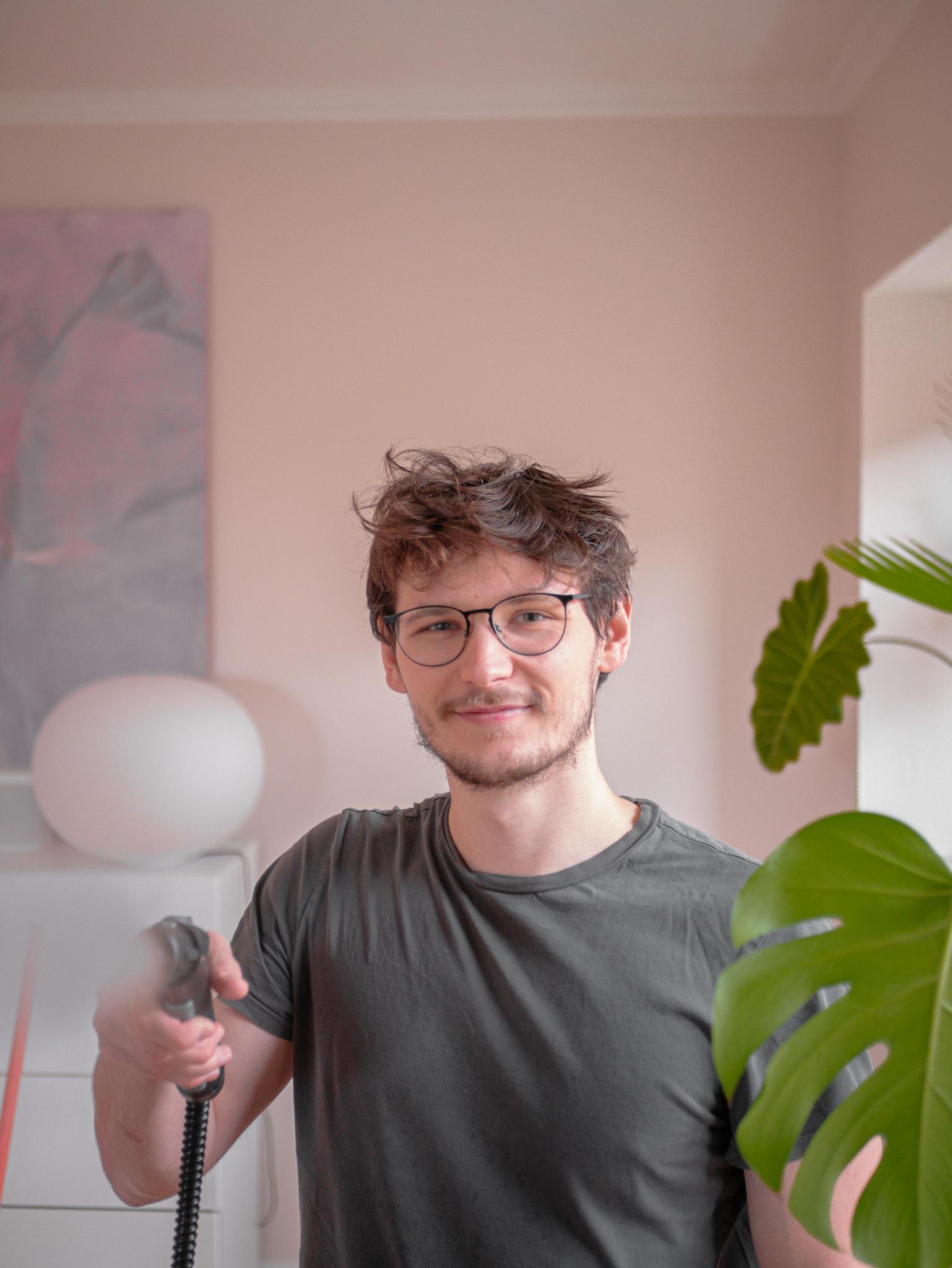 En ung mand med briller står i stuen og holder en støvsuger, omgivet af planter og indretning.