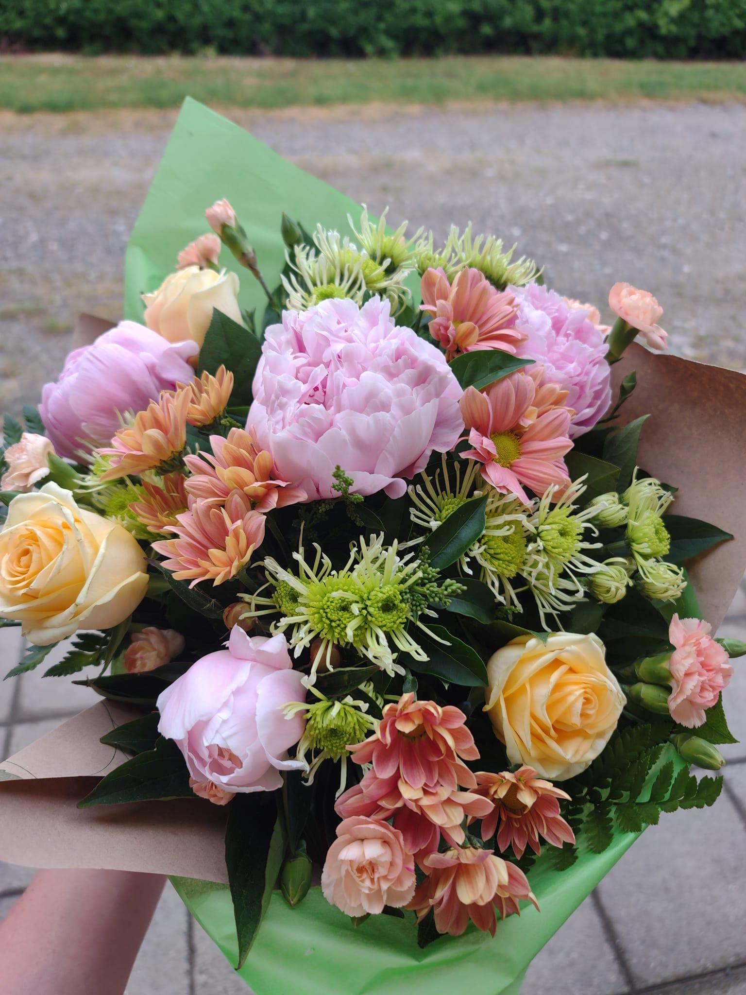 En farverig buket med lyserøde pæoner, orange chrysantemum og gule roser, indpakket i grønt papir.
