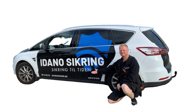 En mand i arbejdstøj står ved siden af en bil med IDANO SIKRING logo og tekst, der reklamerer for sikkerhedstjenester.