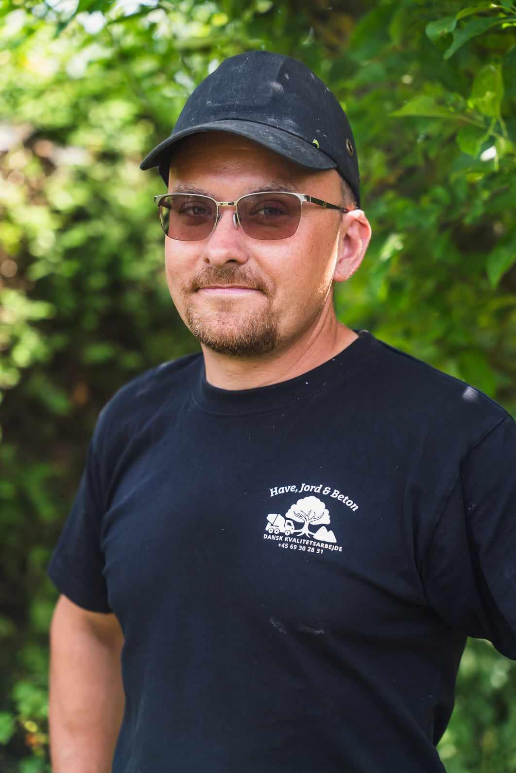 En mand iført solbriller og en sort kasket står foran grønne planter. Han bærer en sort t-shirt med logo.