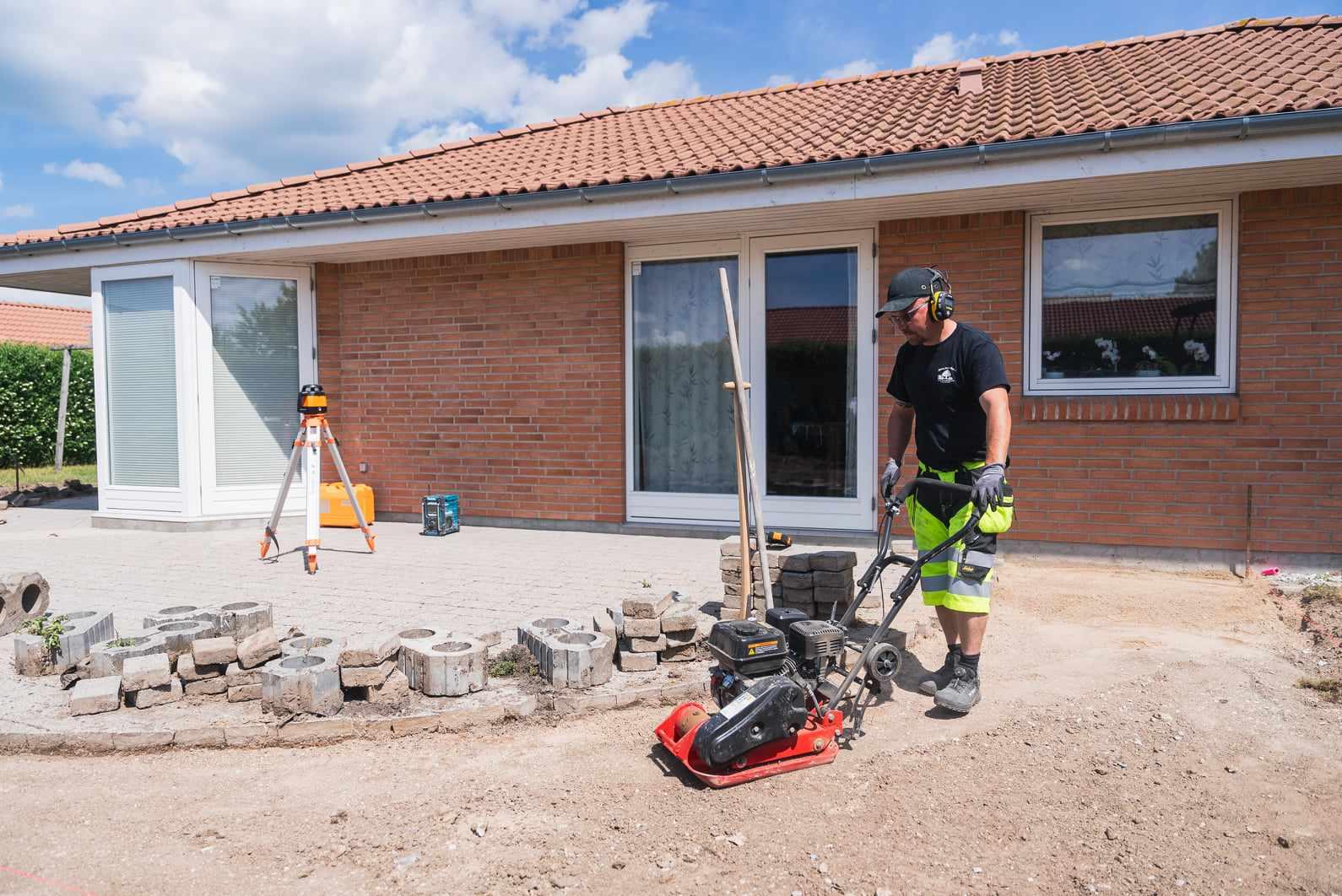 En mand arbejder med en pladevibrator foran et hus med rød murstensvæg og flisebelægning.