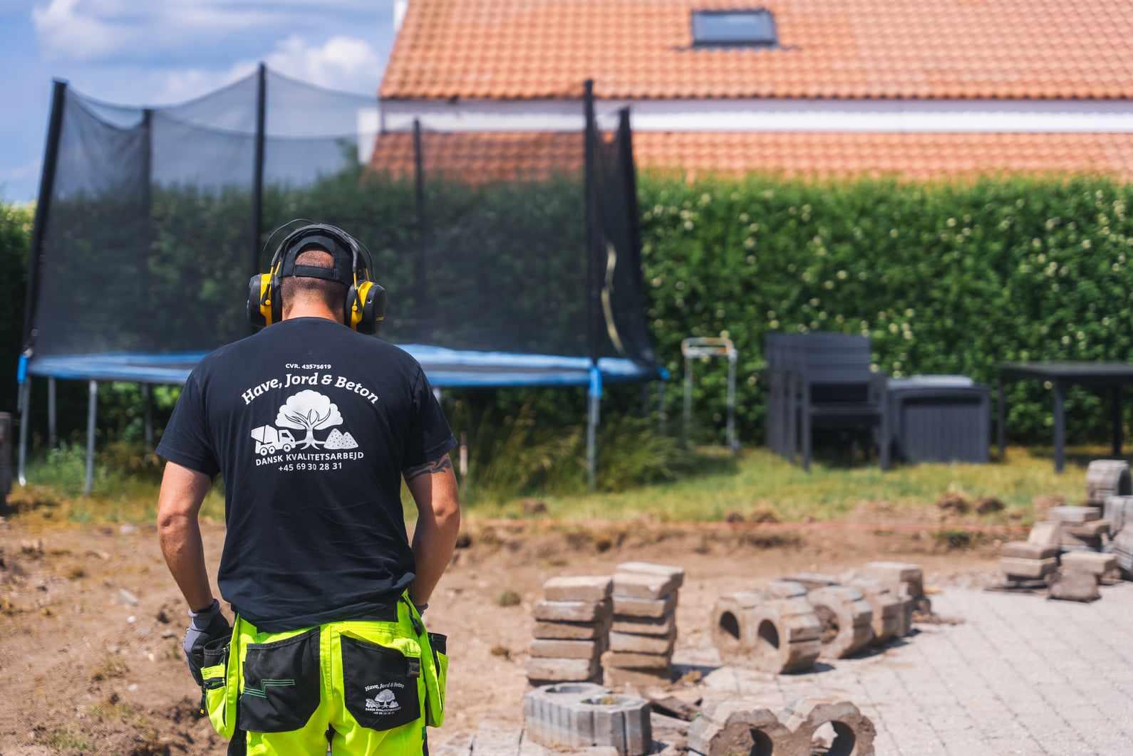 En mand i arbejdstøj står foran en trampolin og en terrasse, mens han arbejder med sten i haven.
