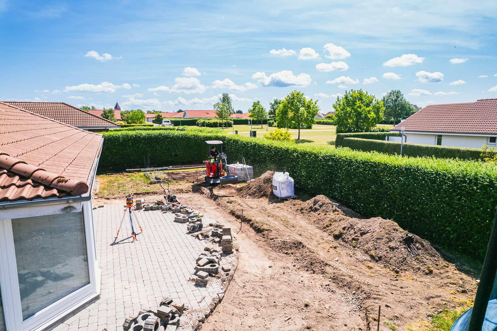 En have med en gravemaskine, der arbejder på en ny belægning, omgivet af grønne buske og et klart blåt himmel.